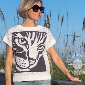 May include: White crochet top featuring a black and white cat face design. The cat's eyes are large and the design is detailed. The top has short sleeves and a relaxed fit.