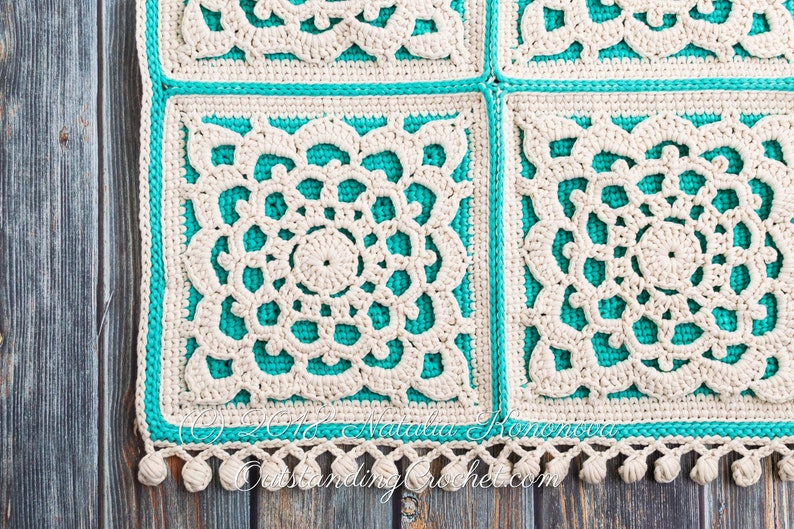 Puede incluir: Una manta de ganchillo con un dise&ntilde;o blanco y turquesa. La manta est&aacute; hecha de cuadrados con un patr&oacute;n floral. La manta tiene un borde de flecos con borlas.