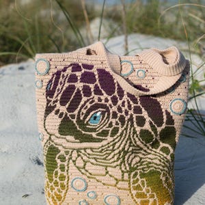 Könnte beinhalten: Beige gehäkelte Tragetasche mit einem Seeschildkröten-Design. Der Kopf und der Panzer der Schildkröte sind in Grün-, Lila- und Gelbtönen gehalten, mit einem blauen Auge. Die Tasche hat einen Schulterriemen und ist mit blauen Kreisen verziert.