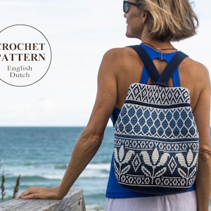 Puede incluir: Una mujer con un top azul y pantalones blancos está de pie en una plataforma de madera con vistas al océano. Lleva gafas de sol y lleva una mochila de crochet azul y blanca con un patrón floral. La mochila tiene correas ajustables y un cierre de cremallera. La imagen incluye el texto "CROCHET PATTERN English Dutch".