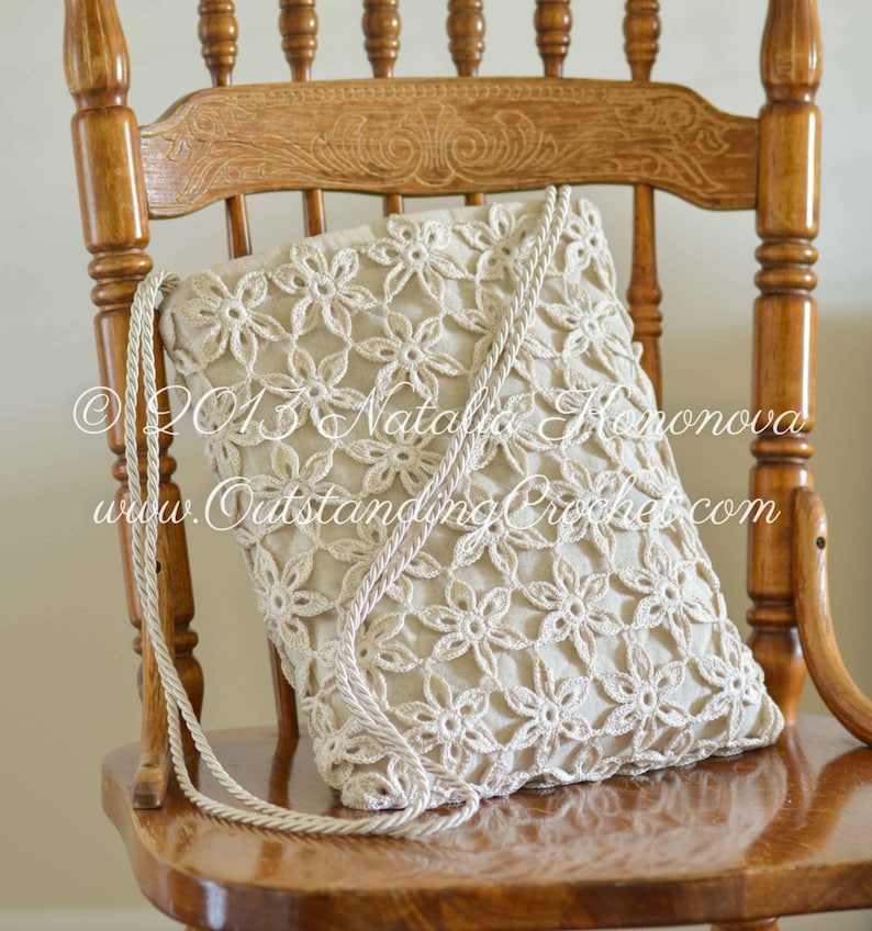 May include: A crocheted bag with a floral design, featuring a beige color and a rope strap. The bag is sitting on a wooden chair.