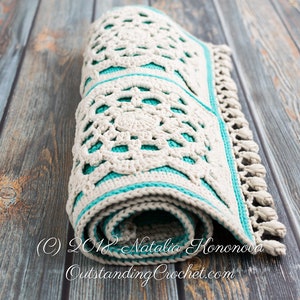 Puede incluir: Una alfombra de crochet enrollada con un dise&ntilde;o blanco y turquesa. La alfombra tiene un borde de flecos y est&aacute; hecha con un patr&oacute;n de puntada delicado.