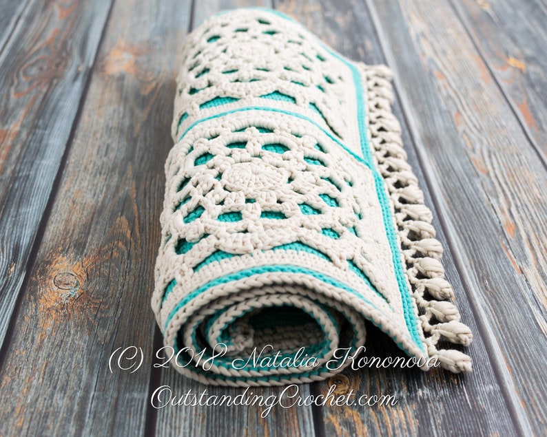 Puede incluir: Una alfombra de crochet enrollada con un dise&ntilde;o blanco y turquesa. La alfombra tiene un borde de flecos y est&aacute; hecha con un patr&oacute;n de puntada delicado.