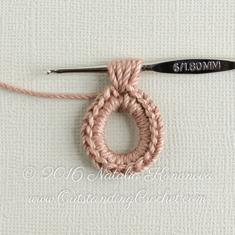 May include: A close-up of a crocheted ring made with a light pink yarn. The ring is being worked on with a 6/1.80 mm crochet hook.