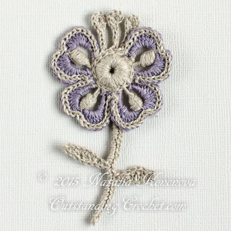 May include: A crocheted flower in a purple and beige colour scheme. The flower has five petals and a centre with a small hole. The flower is attached to a stem with two leaves.