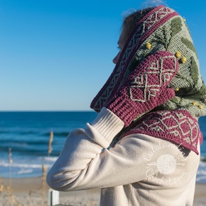 May include: A woman wearing a green and purple crocheted hooded scarf with matching fingerless gloves. The scarf has a geometric pattern and the gloves have a Celtic knot design. The woman is standing on a beach with the sea in the background.
