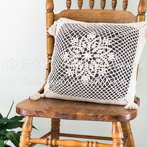 May include: A crocheted cushion with a white lace design on a dark grey background. The cushion has tassels on the corners and is sitting on a wooden chair.