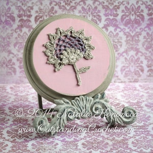 May include: A crocheted flower in shades of beige and purple is displayed on a round, pink and grey wooden plaque. The plaque is on a decorative stand.
