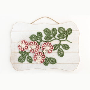 May include: A white wooden sign with a crocheted floral design. The design features pink and cream flowers with green leaves.