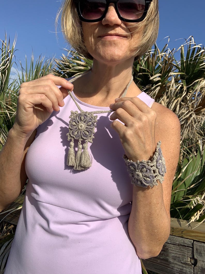 May include: A woman wearing a light purple sleeveless top with a crocheted necklace and bracelet. The necklace has a large, square pendant with a floral design and tassels. The bracelet is a wide band with a similar floral design.