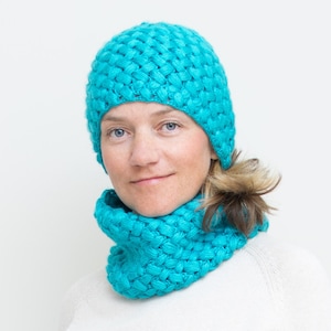 Könnte beinhalten: Eine türkisblaue Strickmütze und ein passender Halsschmuck. Die Mütze ist im Slouchy-Stil mit einer gerippten Textur. Der Halsschmuck ist breit und gemütlich.