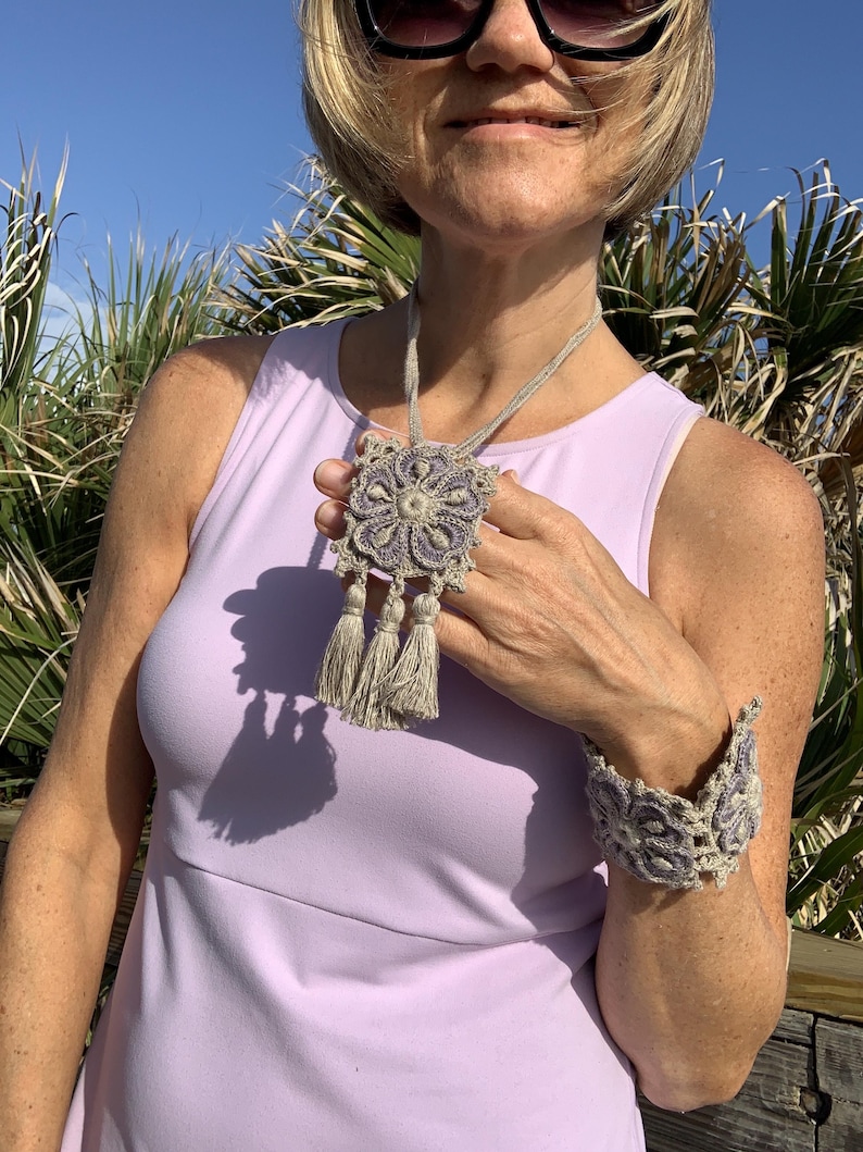May include: A crocheted necklace and bracelet set with a floral design in shades of purple and beige. The necklace features a square pendant with tassels.