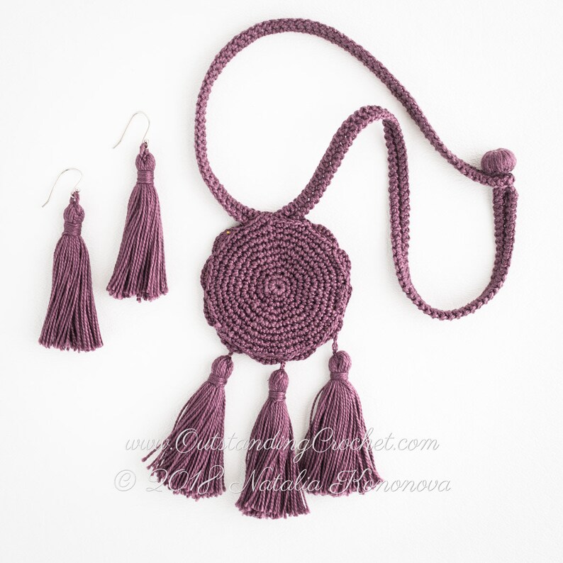 May include: A set of purple crocheted jewelry featuring a round pendant with tassels and matching earrings with tassels. The pendant is made with a spiral pattern and hangs from a long purple cord.