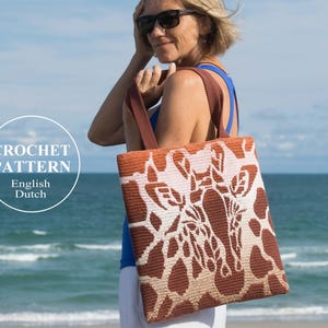 May include: A crochet tote bag with a giraffe pattern in brown and white. The bag has a brown strap and is being held by a person on a beach.