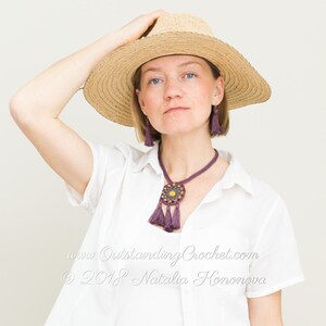 May include: A woman wearing a white shirt and a wide-brimmed straw hat. She is wearing a purple crocheted necklace with a tassel pendant and matching earrings. The necklace features a circular design with a yellow center.