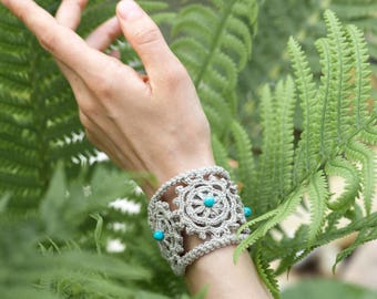May include: A crocheted bracelet with a floral design and turquoise beads. The bracelet is made of a light beige yarn and is worn on a person's wrist.