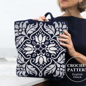 Puede incluir: Una bolsa de mano de crochet con un patrón floral blanco y azul. La bolsa está hecha con un hilo grueso y tiene un asa ancha y resistente. La bolsa está siendo sostenida por una persona frente a un fondo de playa. El texto "CROCHET PATTERN English Dutch" es visible en la esquina inferior derecha de la imagen.
