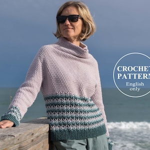 May include: A crochet sweater pattern in English, featuring a pink and green colour scheme. The sweater has a cowl neck and a striped pattern. The model is wearing the sweater and standing on a wooden pier with the ocean in the background.