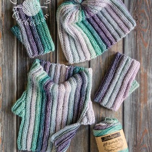 Può includere: Un set fatto a mano composto da cappello, scaldacollo e guanti senza dita nei toni del verde acqua, viola e grigio. È incluso anche un gomitolo di filato abbinato. Il set è esposto su una superficie di legno.