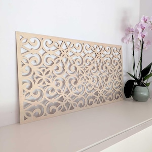 Op de afbeelding: Een lichtgekleurd houten wandkunstpaneel met een herhalend bloemen- en krulwerkontwerp. Het rechthoekige paneel wordt op een wit oppervlak weergegeven. Een orchidee in pot met roze bloemen staat op de achtergrond.