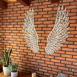 Op de afbeelding: Een paar grote, lichtgekleurde houten wanddecoraties in de vorm van gedetailleerde vogelvleugels met ingewikkelde, lasergesneden veerpatronen. De twee afzonderlijke vleugelsegmenten zijn symmetrisch bevestigd op een roodbruine bakstenen muur.