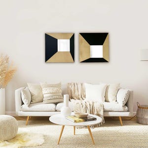 Op de afbeelding: Twee vierkante wandspiegels met een geometrisch ontwerp. Elke spiegel heeft een zwart en goudkleurig frame met een vierkante spiegel in het midden. De spiegels zijn gemonteerd op een witte muur boven een lichtgekleurde bank met decoratieve kussens.