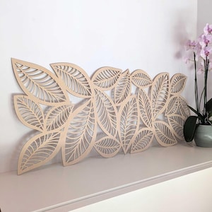 Può includere: Decorazione murale in legno a forma di foglie. Le foglie sono ritagliate, creando un design dettagliato. L'opera d'arte è marrone chiaro e poggia su una superficie bianca. Sullo sfondo è visibile un'orchidea in vaso.