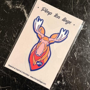 Peut inclure: Un autocollant représentant une tête d'orignal stylisée orange et rose avec des bois blancs et un contour bleu. L'orignal a un cœur bleu sur la poitrine avec le texte blanc MTL. L'autocollant est sur une carte avec le texte Pimp ton linge.