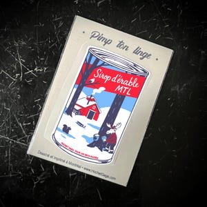 May include: A sticker with a cartoon maple syrup can design, depicting a winter scene with a red cabin, a moose, and a squirrel. The can reads "Sirop d'érable MTL". The text "Pimp ton linge" is above the sticker.