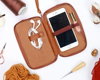 Pode incluir: Estojo de telefone de couro marrom aberto com zíper, alça de pulso e faixa elástica. Dentro, um smartphone branco, fones de ouvido brancos e um tubo de gloss labial são visíveis. O estojo é cercado por materiais de costura e fios.