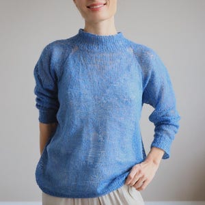 Puede incluir: Un suéter azul claro y semitransparente con cuello alto y mangas largas. El suéter se usa con pantalones beige. La mujer está sonriendo. El suéter parece estar tejido.