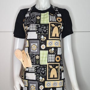 May include: Black apron with a patchwork design featuring bees, flowers, and beehives. The apron has a large pocket on the front and is made of cotton fabric.