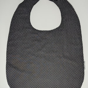 May include: A black and white polka dot bib with a curved bottom and a neck opening. The bib has a hook and loop closure at the neck and is made of a soft fabric. The bib is designed to protect clothing from spills and stains.