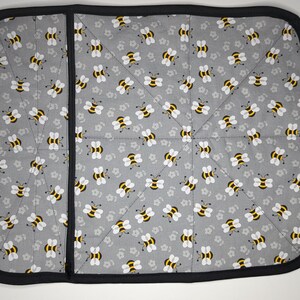 May include: A gray and black pot holder with a bee pattern. The pot holder is made of fabric and has a black border.