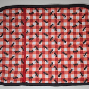 May include: Red and white checkered hot pad with black ants printed on it. The hot pad has a black border and a red zipper.
