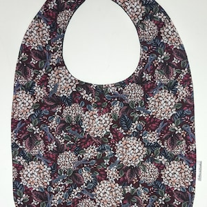 Pode incluir: Um babador bordô com um padrão floral que apresenta flores brancas, folhas verdes e libélulas azuis. O babador tem uma alça de pescoço ajustável preta com fecho de gancho e laço. O babador tem uma etiqueta que diz "La Rue de Cambrai".