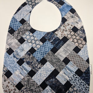 May include: A blue and grey patchwork baby bib with a black border. The bib features a variety of patterns, including floral, geometric, and abstract designs.
