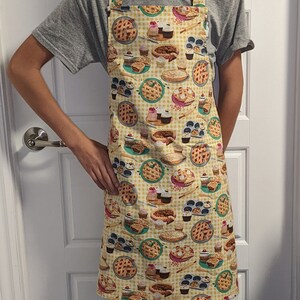 May include: A person wearing a grey t-shirt and a yellow apron with a pattern of pies, cupcakes, and rolling pins. The apron has a checkered pattern and is tied around the waist. The person is wearing blue jeans and yellow socks with a pattern of cars.