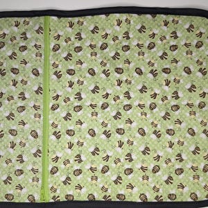 May include: A green and white patterned changing pad with a black border and a green zipper. The pattern features numerous brown and yellow bees. The pad is designed for baby care.