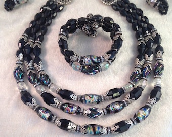 May include: A three-strand black glass bead necklace with silver accents and iridescent glass beads. The set includes matching earrings and a bracelet.