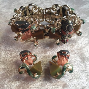 Puede incluir: Un brazalete dorado y unos pendientes a juego presentan un rostro de mujer estilizado con un diseño geométrico verde y marrón.
