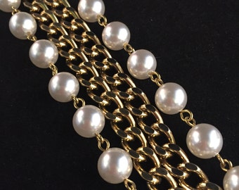 Puede incluir: Primer plano de un collar de cadena dorada con múltiples hilos. Cada hilo presenta grandes perlas redondas blancas. El collar está sobre un fondo negro, resaltando el contraste de colores.