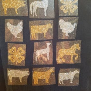 May include: A black fabric background with a grid of 12 squares. Each square contains a glitter-covered silhouette of a farm animal or a four-leaf clover. The animals include a pig, horse, chicken, cow, and sheep. The clover is gold.