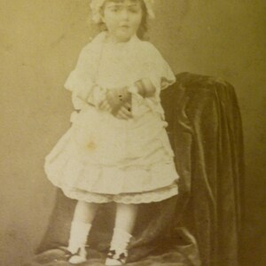 Könnte beinhalten: Ein junges Mädchen in einem weißen Kleid und einer weißen Haube sitzt auf einem Stuhl. Das Foto ist in Sepia-Tönen.