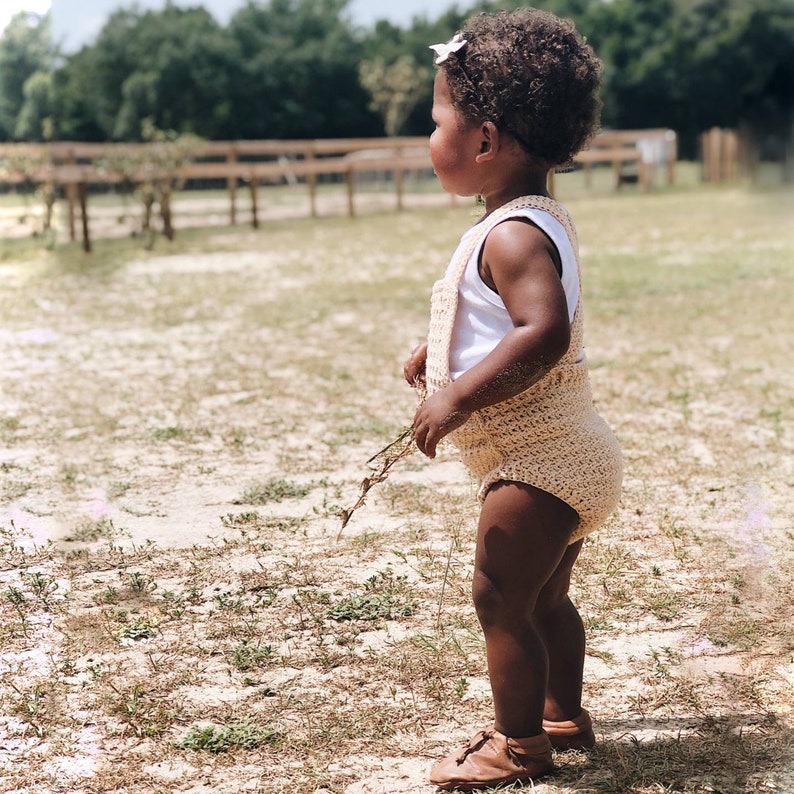 May include: A young child wearing a light brown crocheted romper and brown leather shoes stands in a grassy field. The child is looking to the right of the frame.