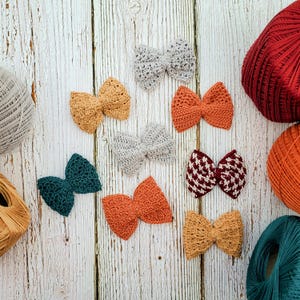 May include: Assortment of handmade crocheted bows in various colors, including teal, orange, red, and beige. The bows are displayed on a white wooden surface.