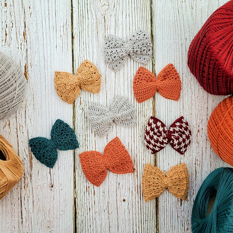 May include: Assortment of handmade crocheted bows in various colors, including teal, orange, red, and beige. The bows are displayed on a white wooden surface.