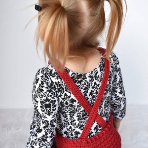 May include: A red crocheted overall with criss-cross straps worn over a black and white patterned shirt.
