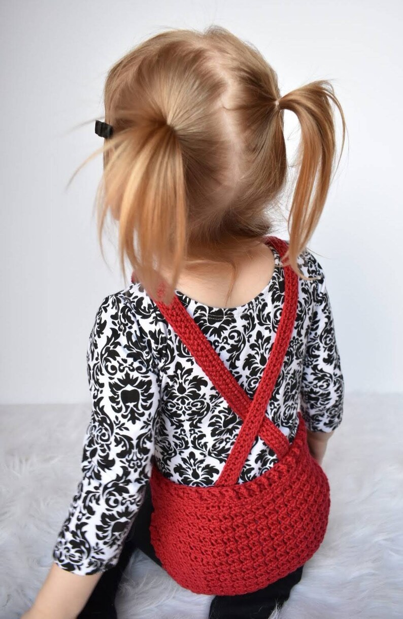 May include: A red crocheted overall with criss-cross straps worn over a black and white patterned shirt.