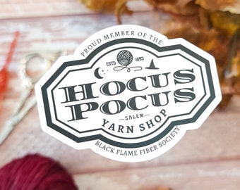 May include: A white sticker with black text that reads "Proud Member of the Hocus Pocus -Salem- Yarn Shop Black Flame Fiber Society" with a black and white illustration of a ball of yarn, a crescent moon, and a witch's hat.